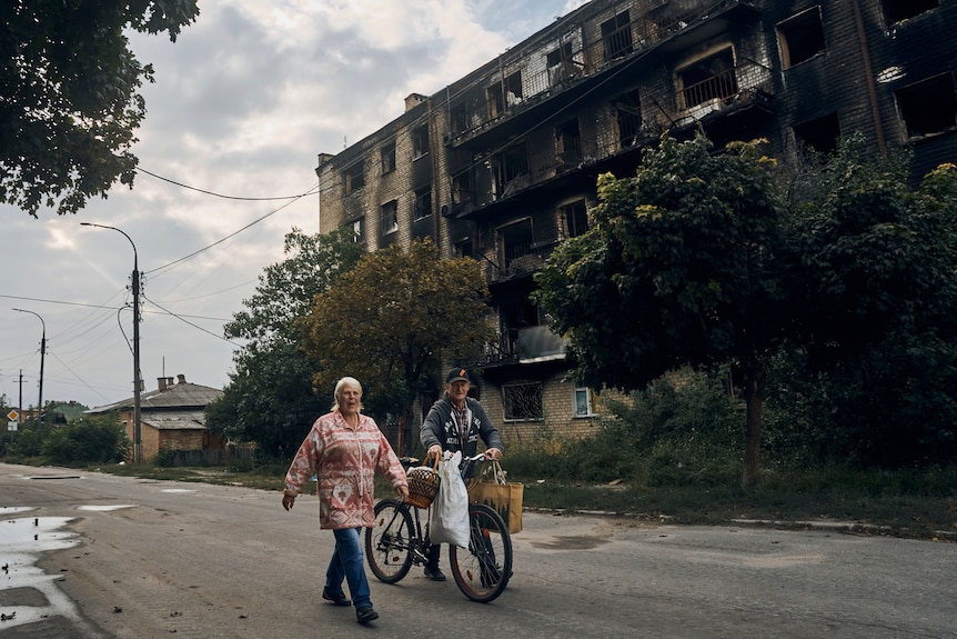 A woman in a bright jacket and a man pushing a bike walk down a road lined with damaged buildings