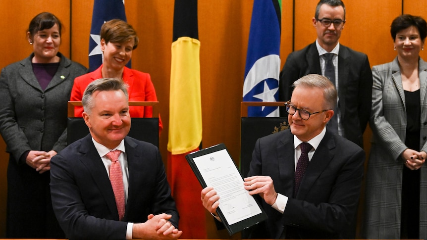 Anthony Albanese and Chris Bowen sit behind a table, as Albanese holds up a document. Four people stand behind them.