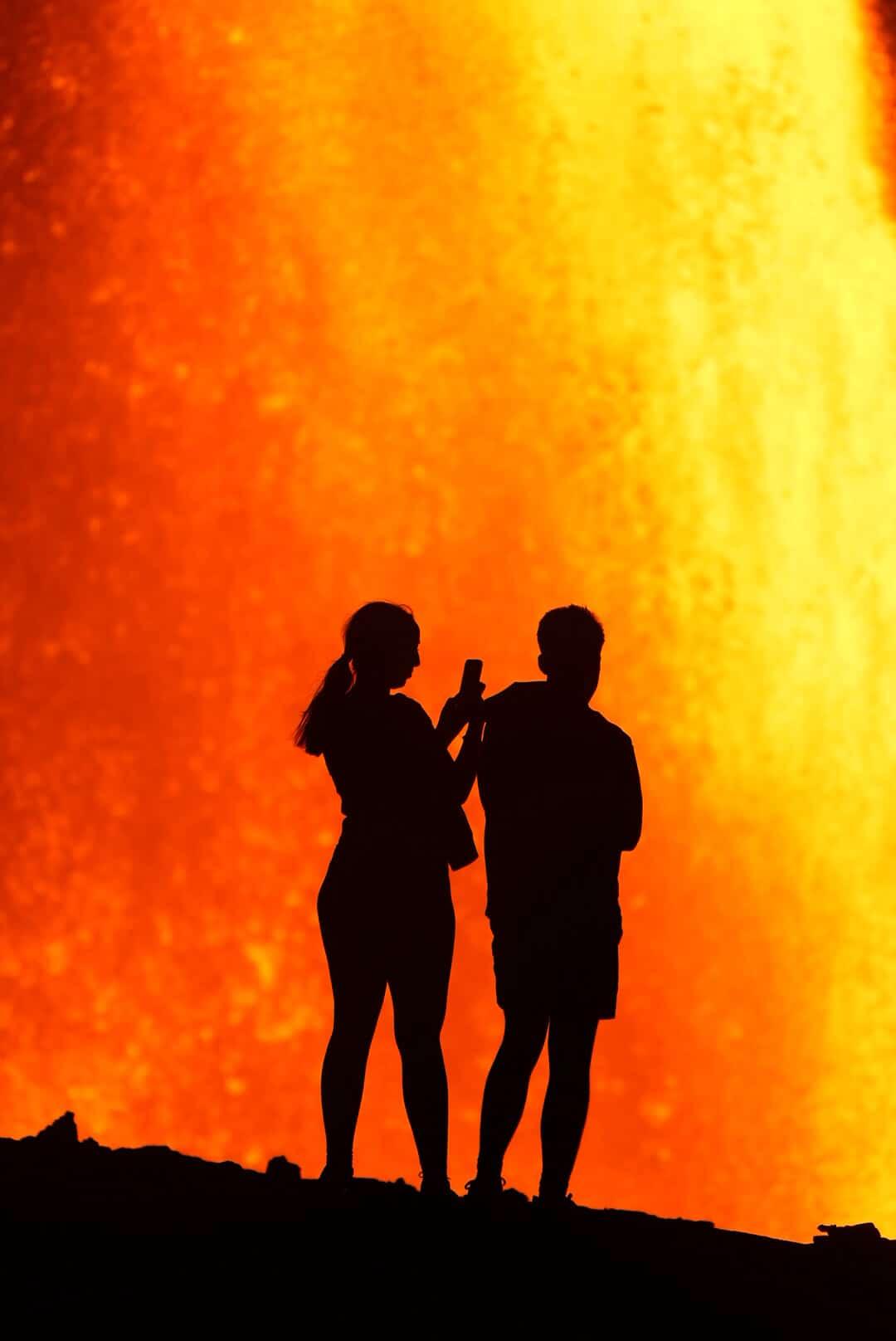 Uma silhueta de duas pessoas em pé com uma fonte de lava laranja brilhante na frente delas.