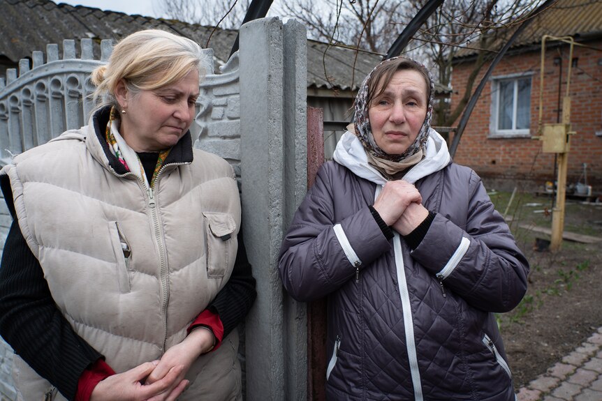 A woman with tears in her eyes clasps her hands at a gate, while a woman in a puffer vest stands next to her