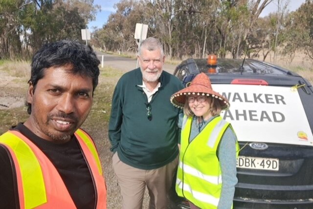 A man in a high vis vest stands with a man and woman in front of a car that has a walker ahead sign 