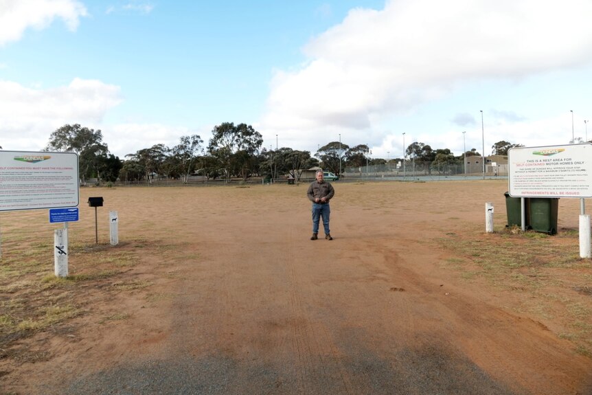 A man wearing blue denim jeans and boots stands in the driveway of an empty caravan park.