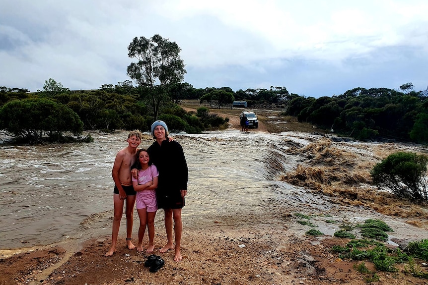 Three people standing in front of a fast-flowing creek, with a vehicle and another person in the background on the other side.