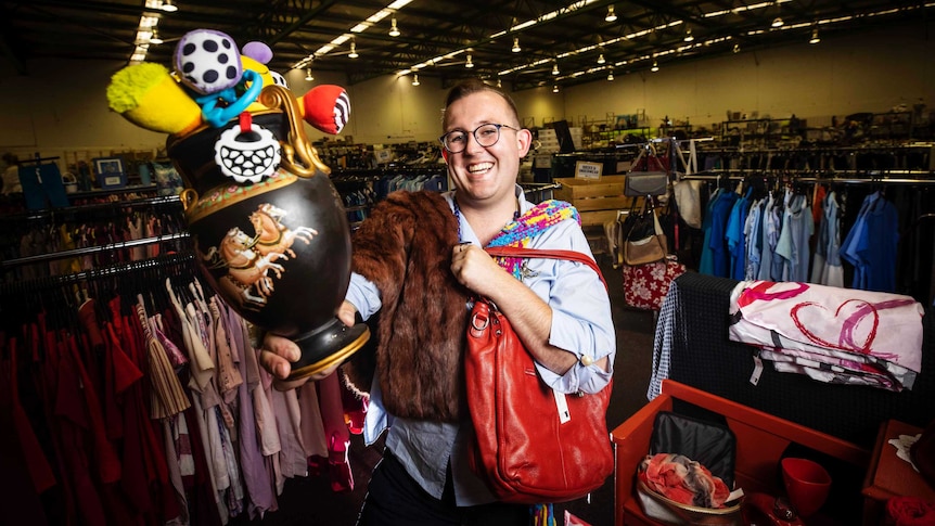 A smiling man in glasses holds and wears a variety of objects in a charity shop.
