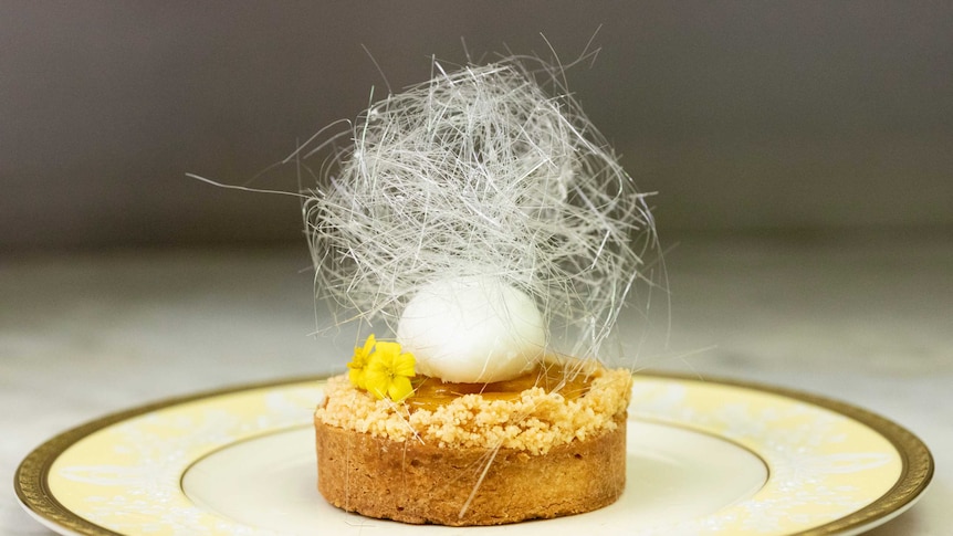 A round tart with a crusting on top, a scoop of white creme fraiche with spun sugar and yellow flowers on top