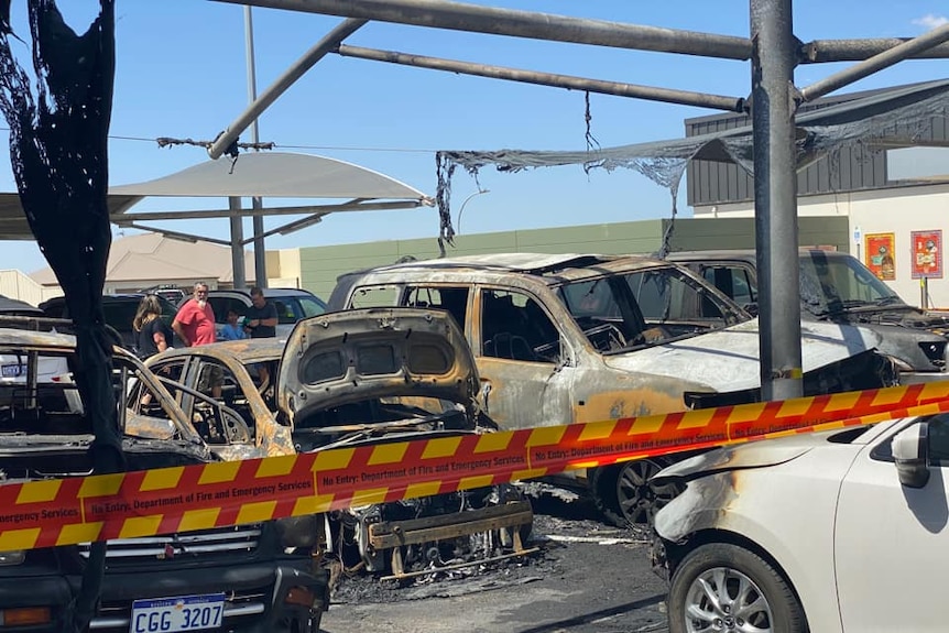 Five cars in a shopping centre are black and destroyed by fire. Shade clothes are melted around the car, three people watch on.