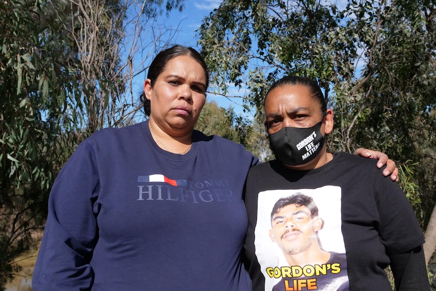 Two Aboriginal women stand together on a riverbank. The woman on the right has a picture of her son on her black T-shirt