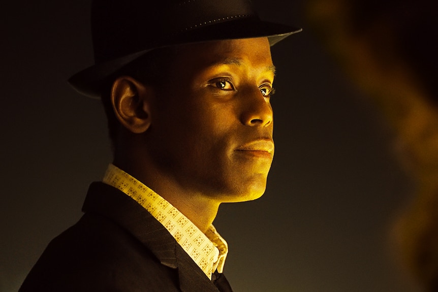 A close up of a black man wearing a black suit and fedora, whose face is lit in warm yellow light looks at someone off camera.