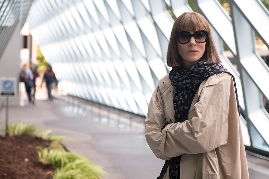 A woman with brown bob, dark sunglasses and trench coat stands in bright walkway corridor near geometric windows.