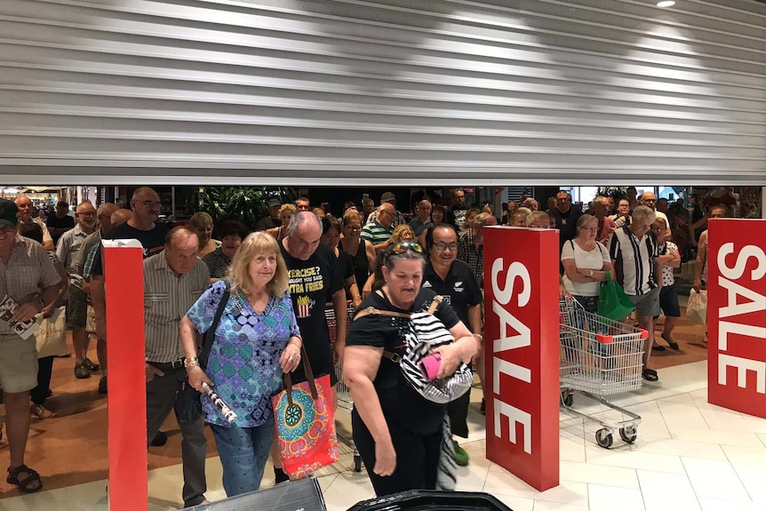 Shoppers walk under a roller door to get into a shopping centre with sales signs.