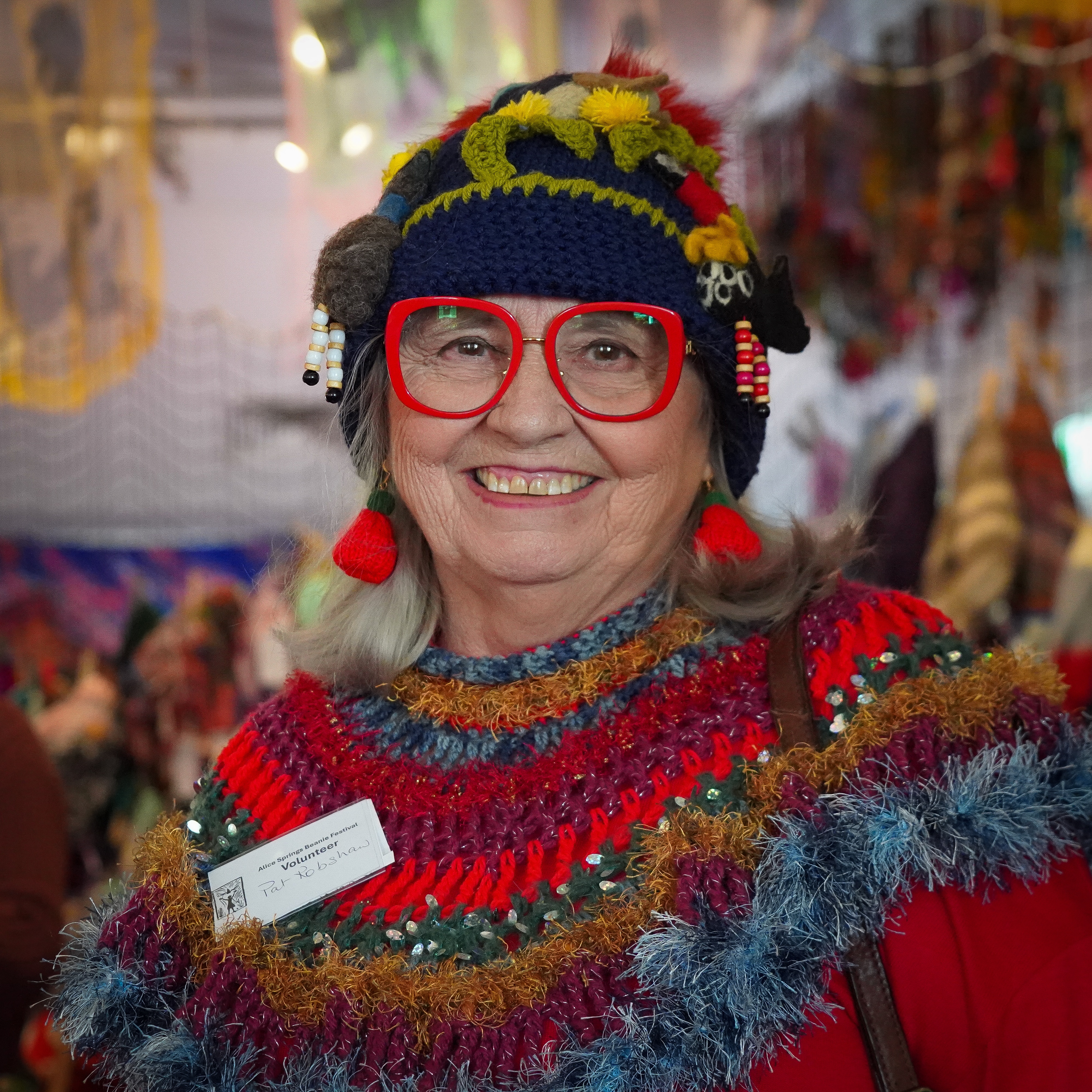 An event volunteer wears red-framed glasses and dangling earrings, a colourful beanie is on her head.