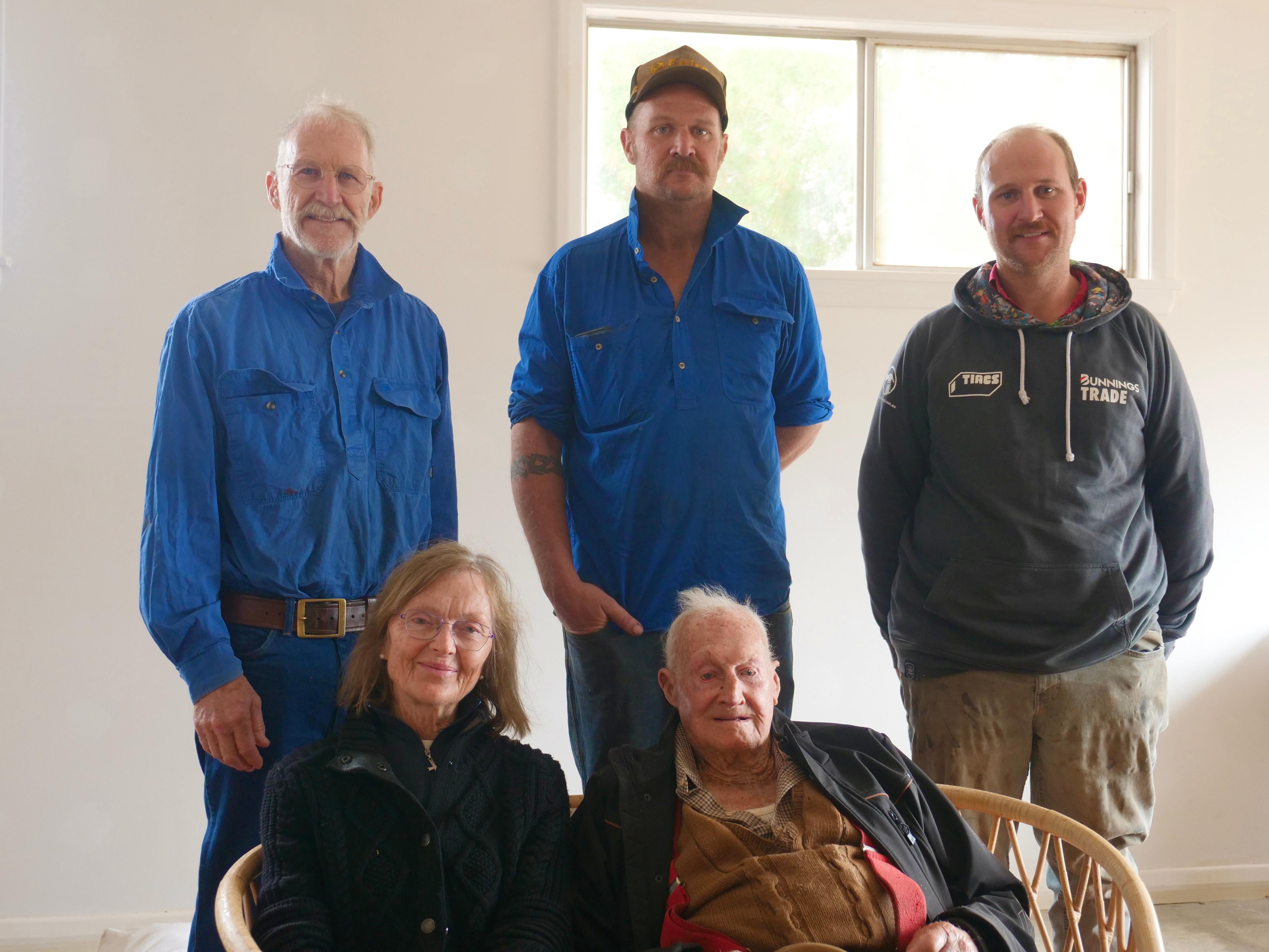 three men standing, two with blue shirts and one with a grey jumper, a lady with glasses sitting next to an old man