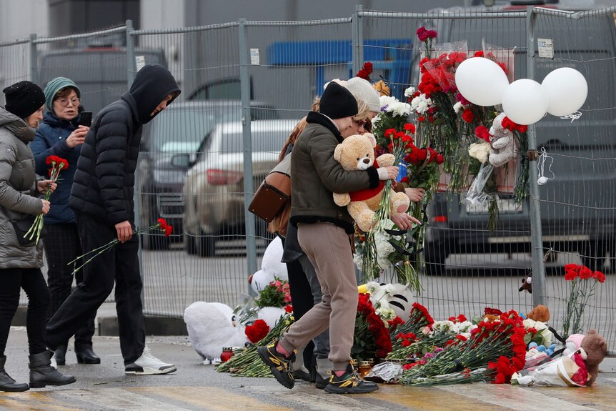 People lay read flowers and teddy bears by a fence.