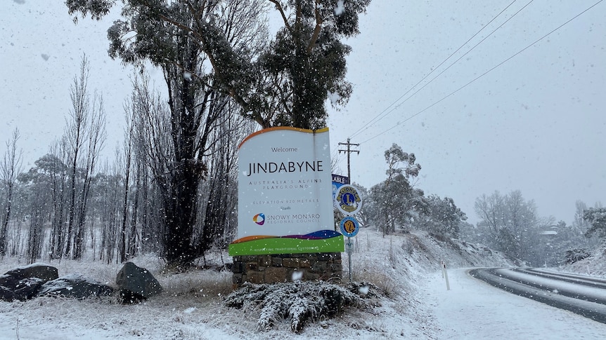 A Jindabyne town sign surrounded by snow as snow falls in the foreground.