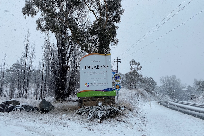 A Jindabyne town sign surrounded by snow as snow falls in the foreground.