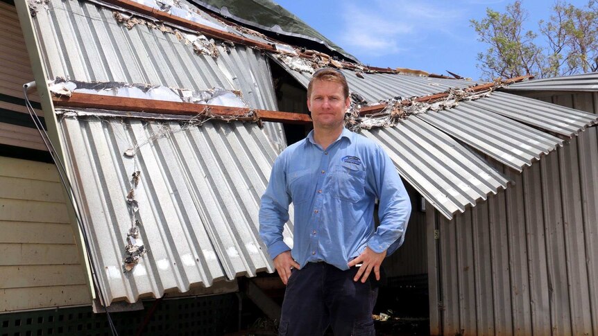 A man stands in front of his home, which has had the roof ripped off.