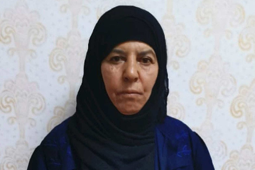 A woman wearing a head scarf stands in front of a wall.