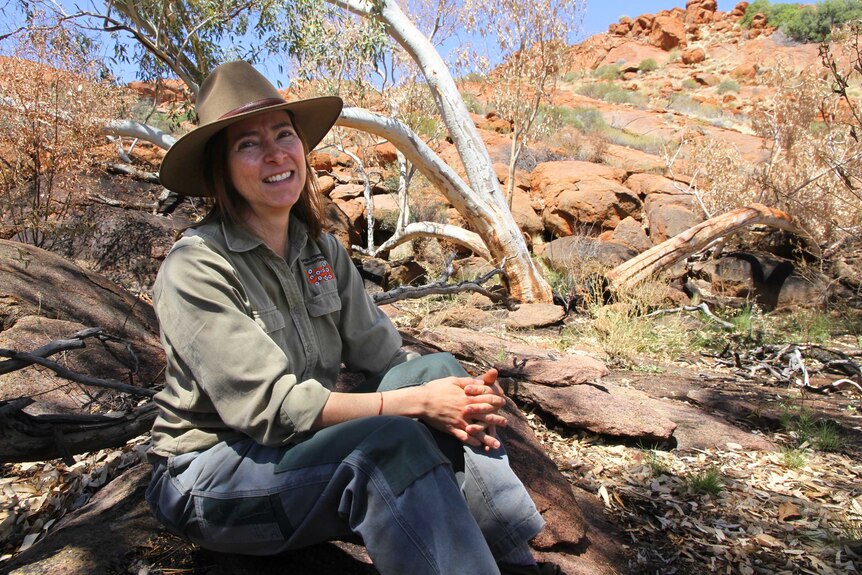 Woman in a hat sitting on a rock in outback scrub.
