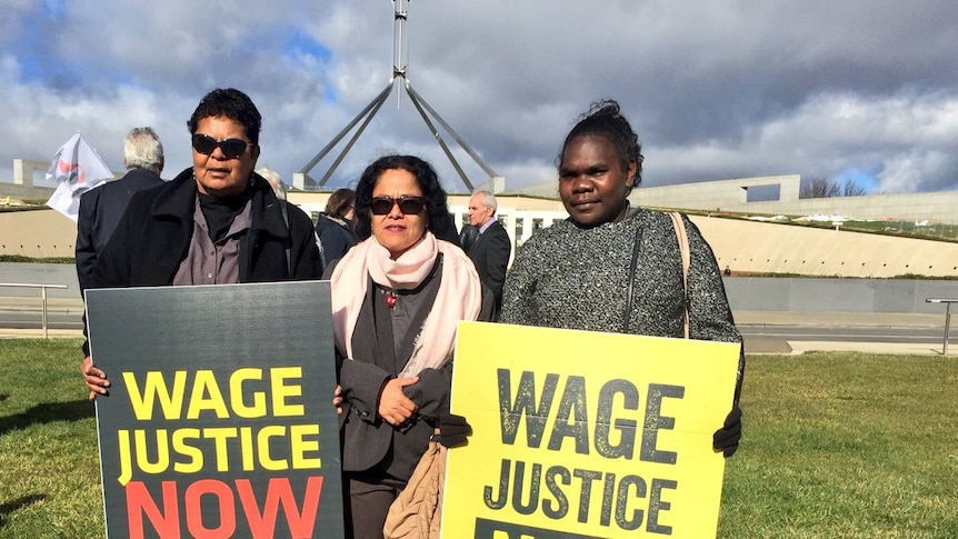 Three women stand with signs saying wage justice now with parliament house in the background.
