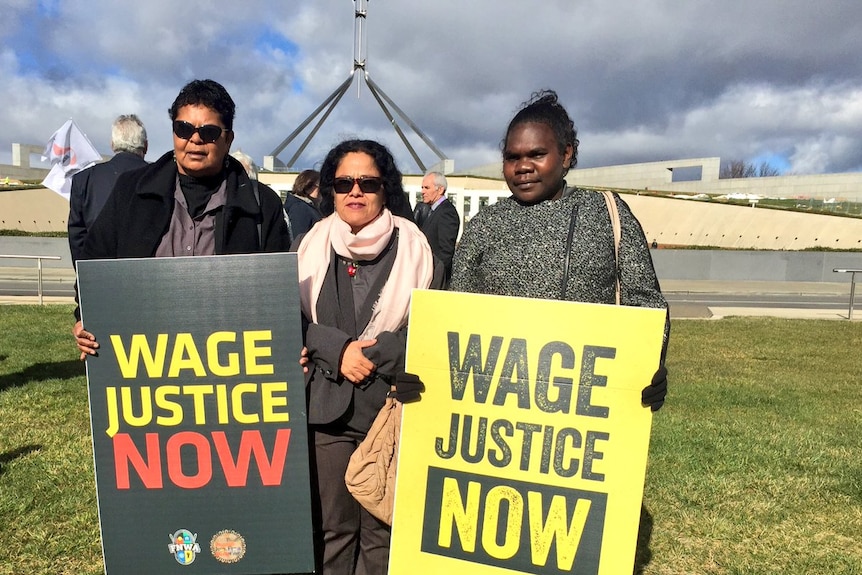 Three women stand with signs saying wage justice now with parliament house in the background.