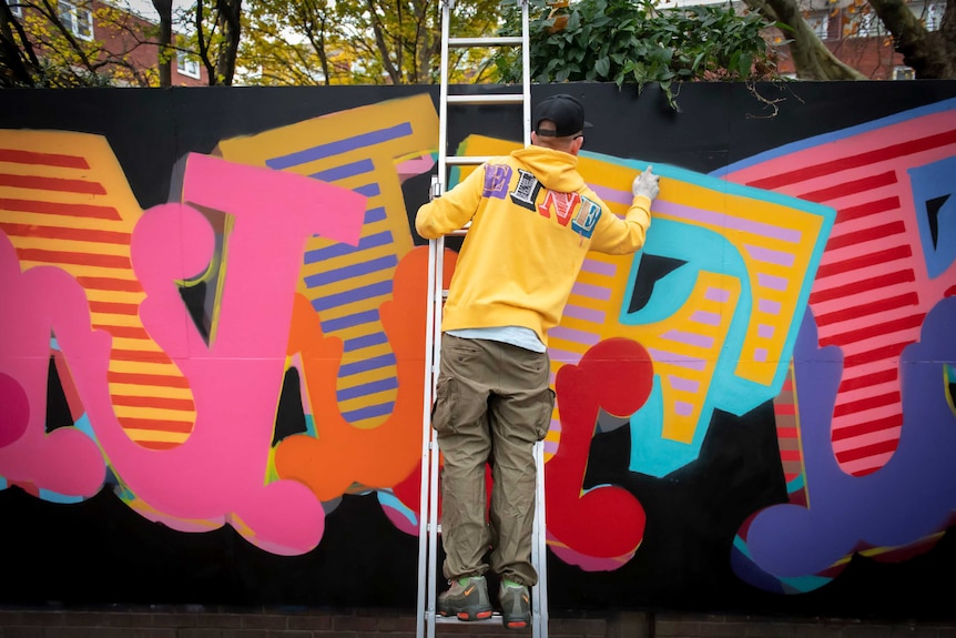 The back of a man in bright yellow hoodie, pants and sneakers, on a ladder spray-painting letters on a brightly painted wall.