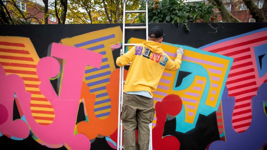 The back of a man in bright yellow hoodie, pants and sneakers, on a ladder spray-painting letters on a brightly painted wall.