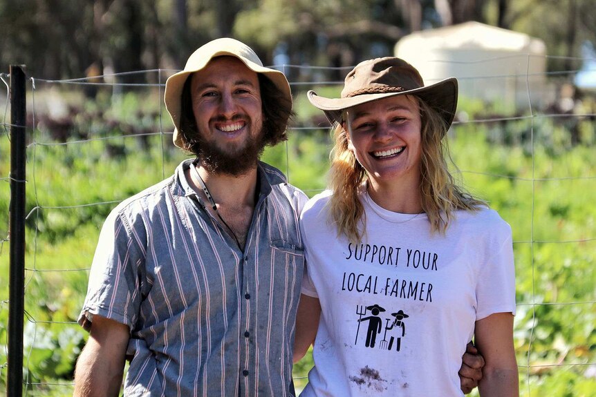 A smiling couple wearing farmers' hats stand together in front of a wire fence.