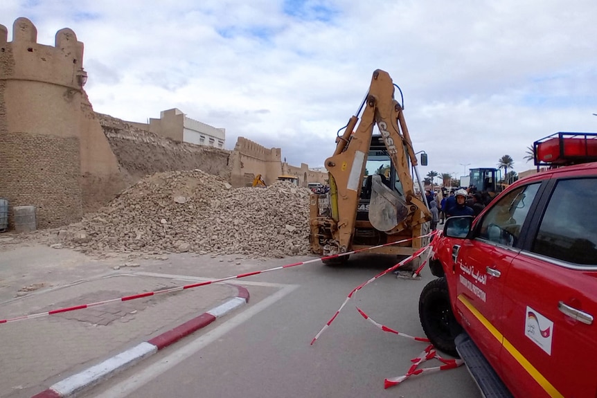 The side of a castle wall collapsed with gravel mounds in front and an excavator and red police car
