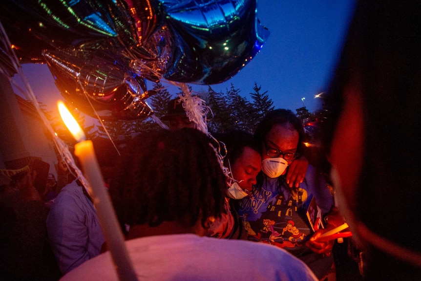 A masked woman and a man hug each other among candles and balloons at a memorial.