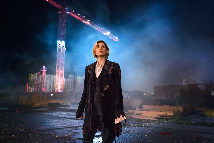 A woman in a tattered coat stands in front of a crane