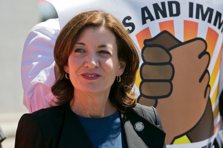 A woman in a suit jacket grins as she looks off camera while standing in front of a sign with cartoon hands shaking.