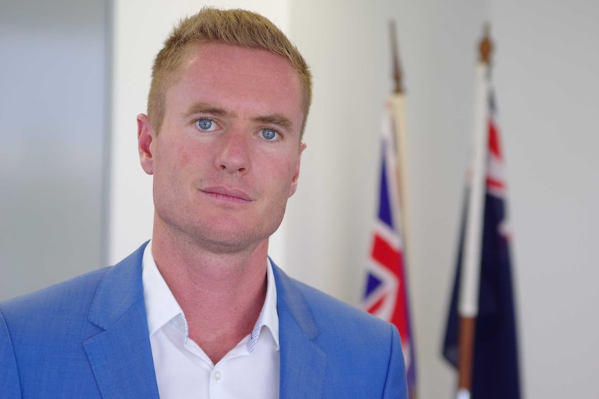 A head and shoulders shot of Albert Jacob in a blue jacket and white shirt with out-of-focus flags in the background.