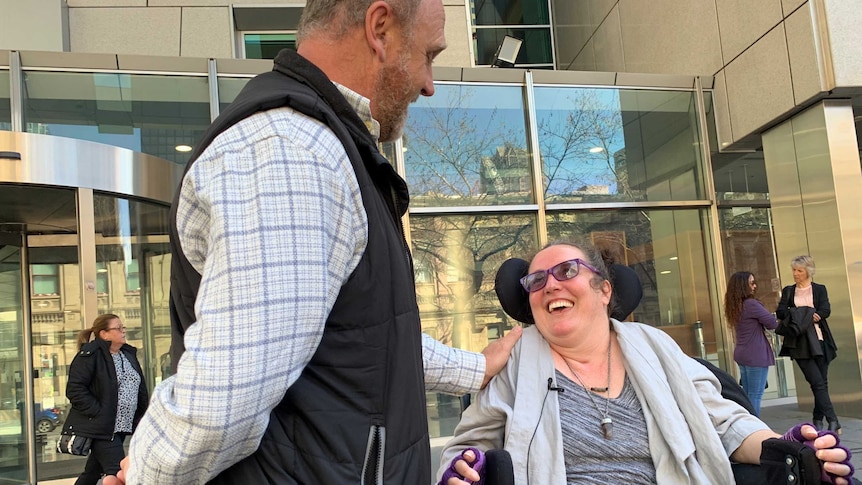 A woman in a wheelchair smiles up at man who has his hand on her shoulder.