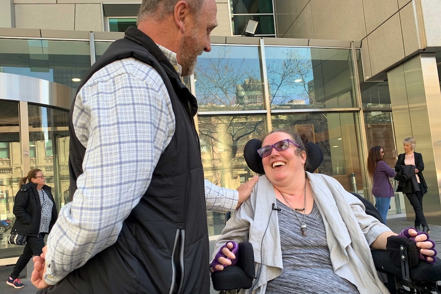 A woman in a wheelchair smiles up at man who has his hand on her shoulder.