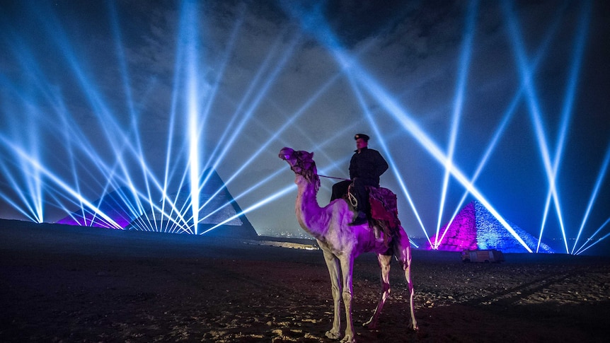 An Egyptian policeman sits on a camel in front of the pyramids and a laser beam show.