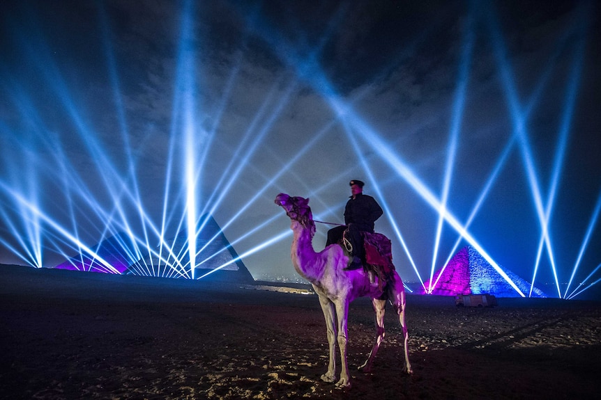 An Egyptian policeman sits on a camel in front of the pyramids and a laser beam show.