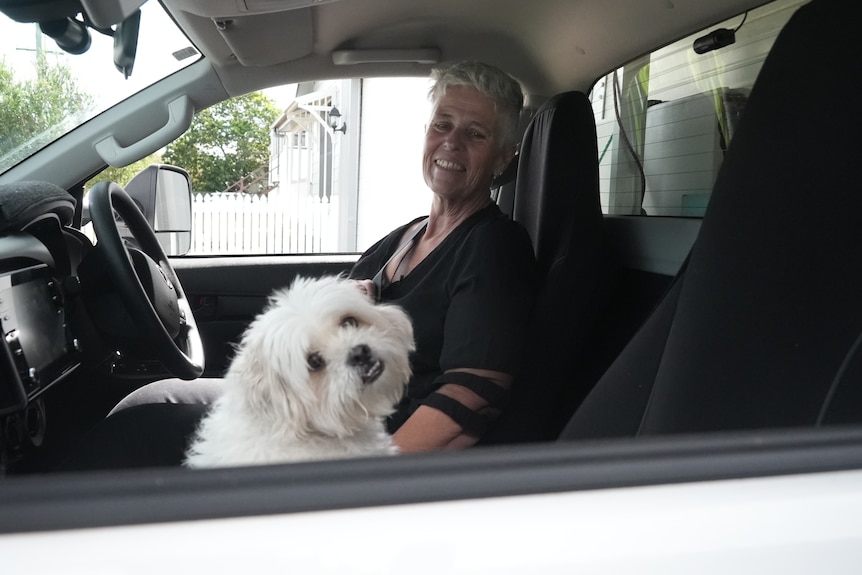 A woman with short grey hair sits behind the wheel of a campervan next to a small white dog