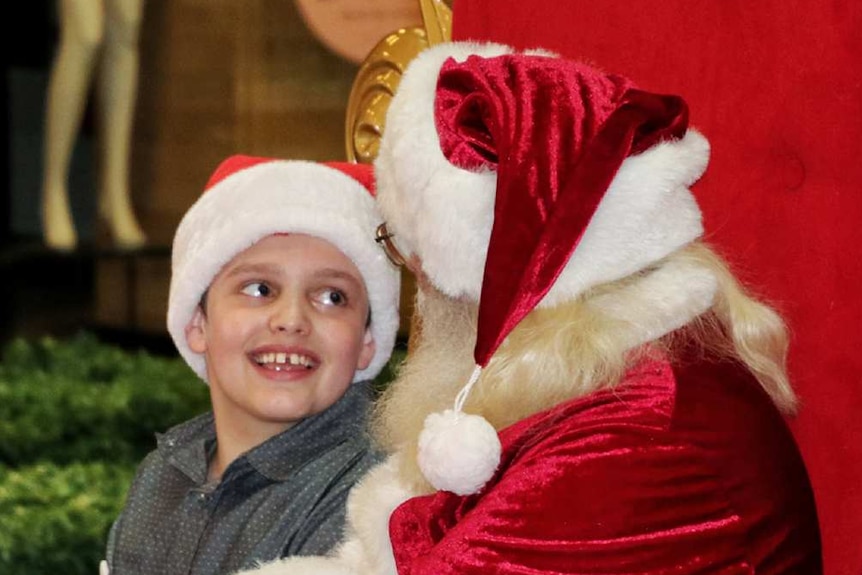A boy in a grey shirt sits on Santa's lap on a big gold and red chair.