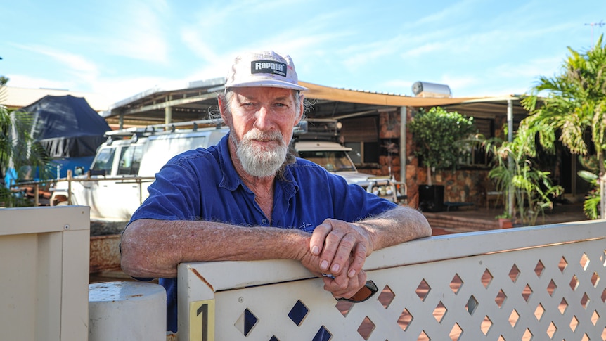 An older man with grey beard stands in front of his house, leans on the white, trellised front gate. 