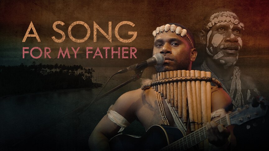 A man wearing traditional garb has panpipes around his neck and a microphone in front of him. Another looks over his shoulder.