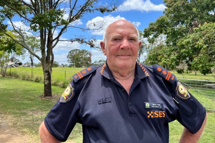 A man in an SES uniform looks at the camera with his hands on his hips, green paddock behind