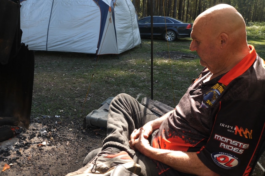 A man sits in a camp chair by his camp fire sun outlining his face and bald head.