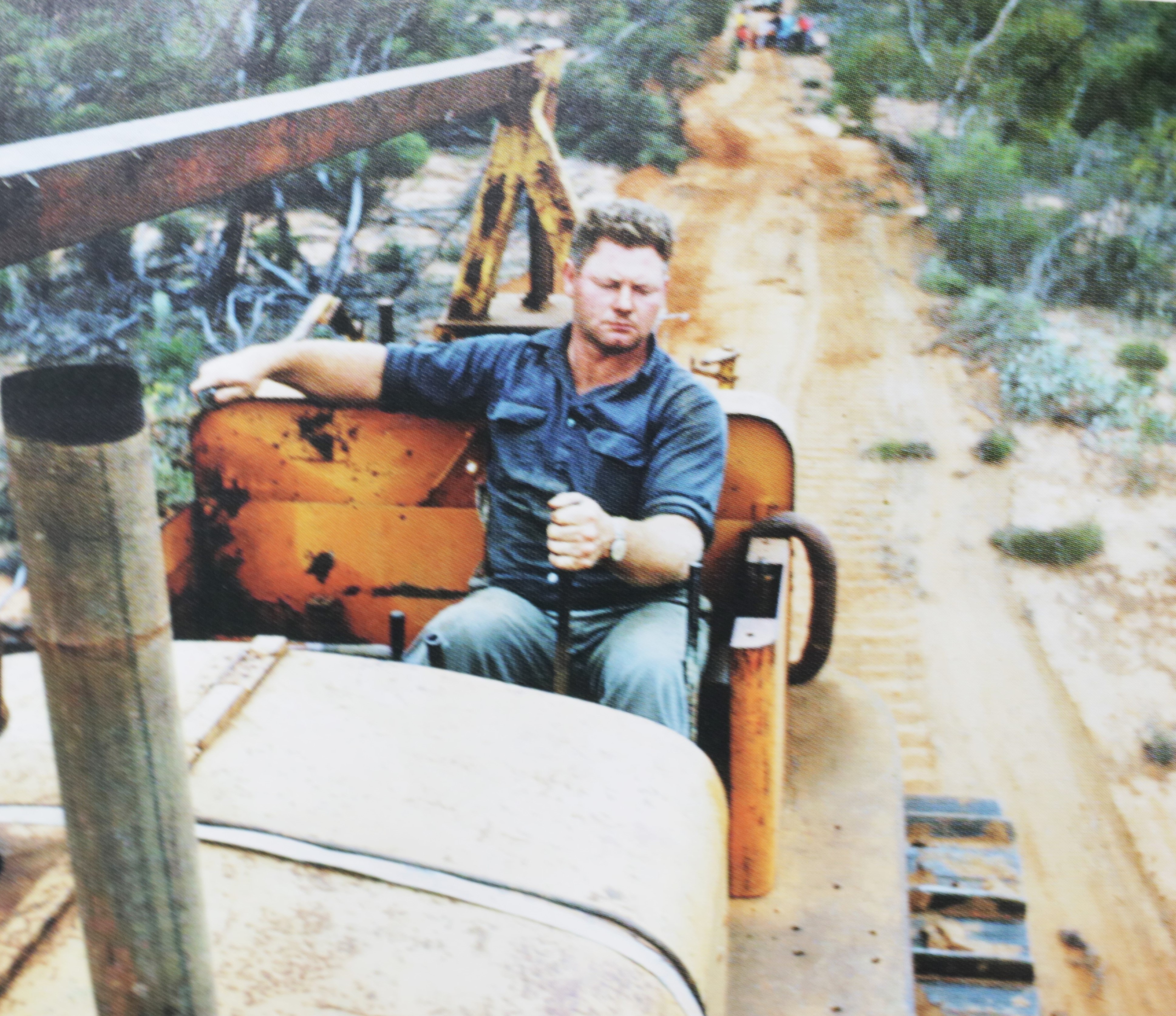 man in blue work short driving cab;ess bulldozer on sandy road, scrub on raodside