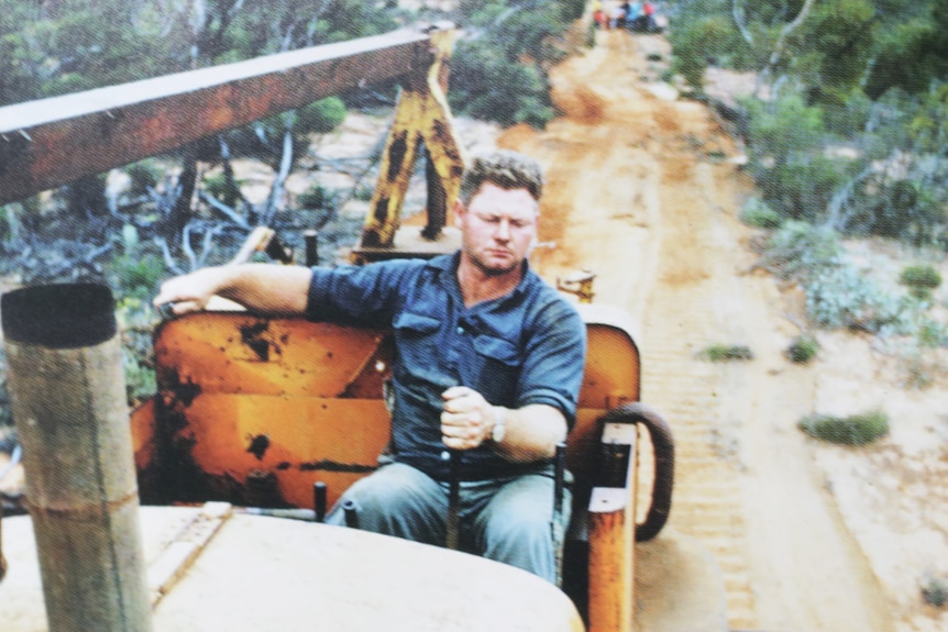 man in blue work short driving cab;ess bulldozer on sandy road, scrub on raodside