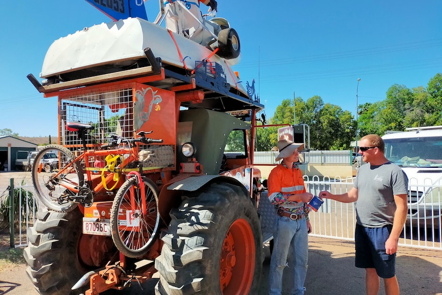 Young man in bright orange shirt stands next to tractor holding out donation tin as a man puts money in it