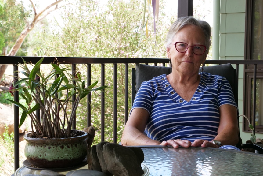 A woman in her 60s in red glasses and a blue striped top sits to the right of a glass outdoor table. She has a serious face.