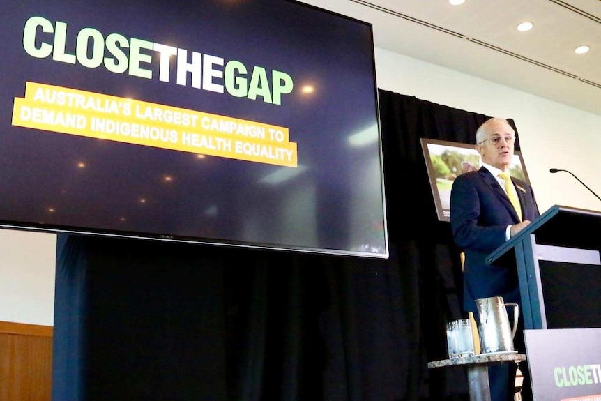 Malcolm Turnbull speaks at a podium, with the Closing the Gap projected onto a screen next to him.