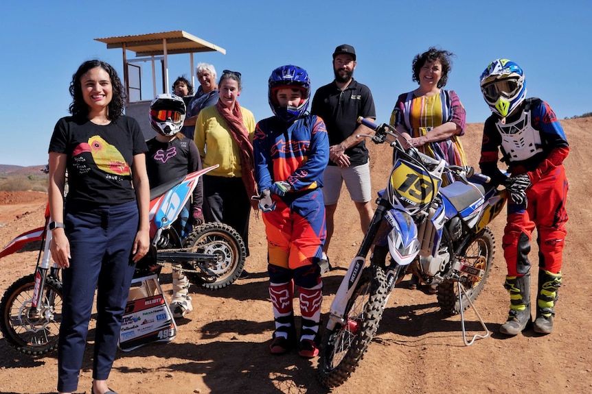 Group of people standing with kids besides dirt bikes on red dirt.