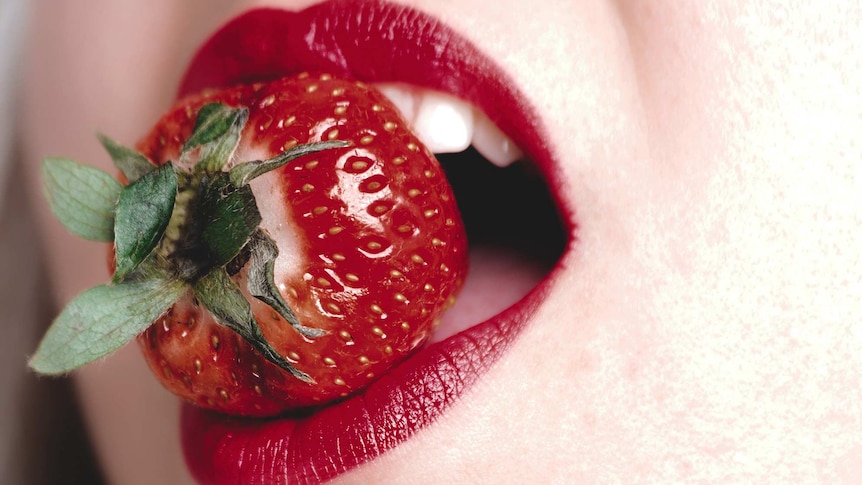 Woman wearing red lipstick with a fresh strawberry in her mouth.