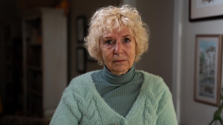 A white-haired woman in blue cardigan holds a tea-cup in both hands.